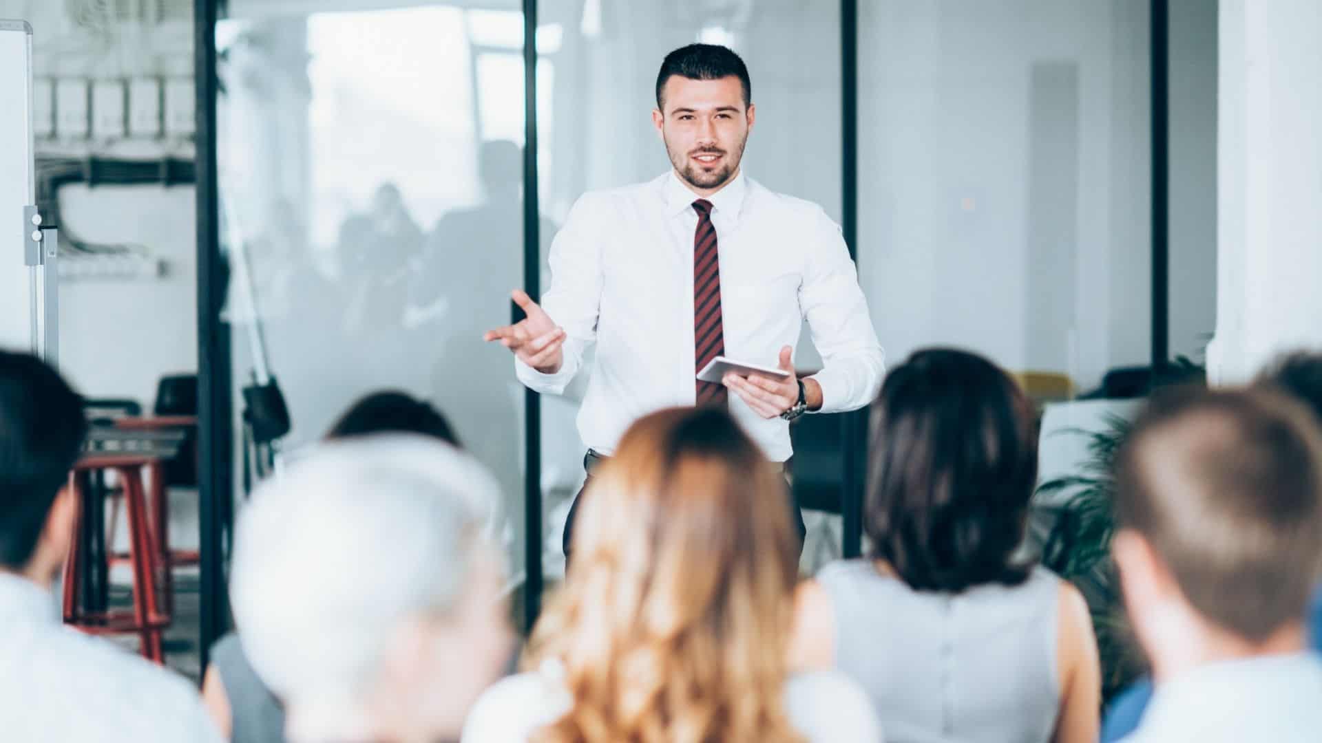 A man in a white shirt and tie discusses cybersecurity services in Contra Costa County, speaking confidently to a seated audience in a bright, modern office. Holding a tablet, he gestures as he talks, capturing the attendees' focus with insights on network security solutions.