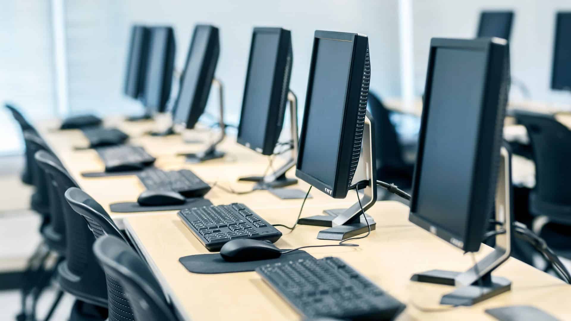 A row of identical desktop computers with monitors, keyboards, and mice are arranged on a long desk in a bright, modern computer lab. Offering top-notch IT support in Contra Costa County, the space features black chairs tucked under the desk as natural light streams through large windows.