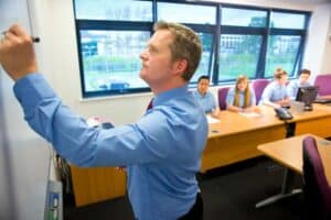 A man in a blue shirt writes about cybersecurity Contra Costa County on a whiteboard at the front of a classroom, while four students seated at desks watch and take notes. Large windows let in natural light.
