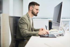 A man with glasses and a beard works at a desk, using a graphic tablet and stylus while looking at a large computer monitor in a modern office—highlighting the need for managed IT Services in Contra Costa County, CA.