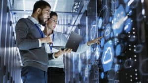 Two IT professionals stand in a server room, examining data on a laptop and interacting with a large digital touchscreen displaying cloud icons—showcasing managed IT services in Contra Costa County, CA.