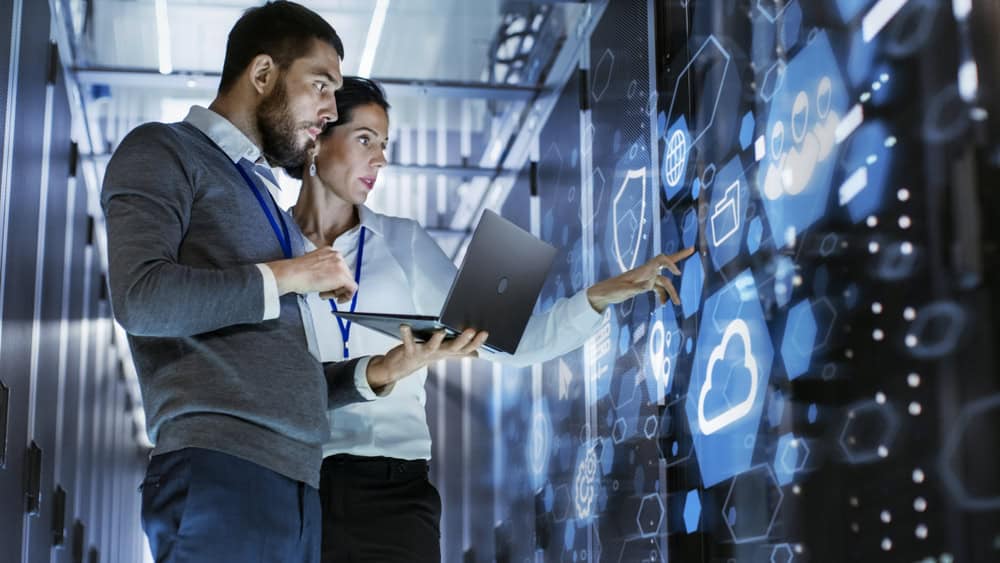 Two IT professionals stand in a server room, examining data on a laptop and interacting with a large digital touchscreen displaying cloud icons—showcasing managed IT services in Contra Costa County, CA.