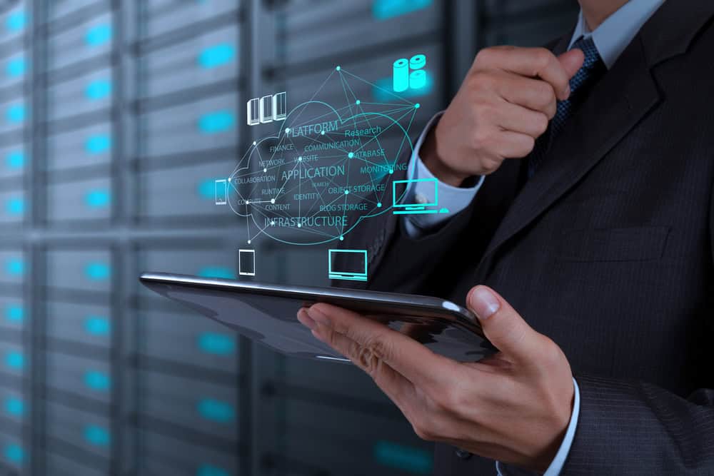 A person in a suit holds a tablet with a virtual cloud computing diagram hovering above it, displaying terms like "platform" and "infrastructure," illustrating managed IT services Contra Costa County in a secure server room setting.