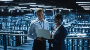 Two IT professionals, a man and a woman, stand in a large, modern data center filled with server racks, smiling and looking at a laptop together—showcasing top-notch cybersecurity Contra Costa County under bright overhead lights.