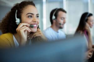 A smiling woman wearing a headset sits at a desk, speaking into a microphone, with two colleagues in headsets working beside her in a bright office focused on managed IT services Contra Costa County.